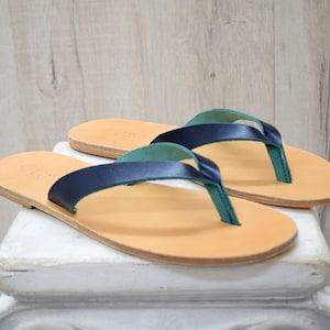Peut inclure: Une paire de tongs en cuir avec des lani&egrave;res bleu marine et des lani&egrave;res de thong vert d'eau. Les sandales ont une semelle marron clair et sont pr&eacute;sent&eacute;es sur une surface blanche sur un fond en bois clair. Chaussures d'&eacute;t&eacute;.