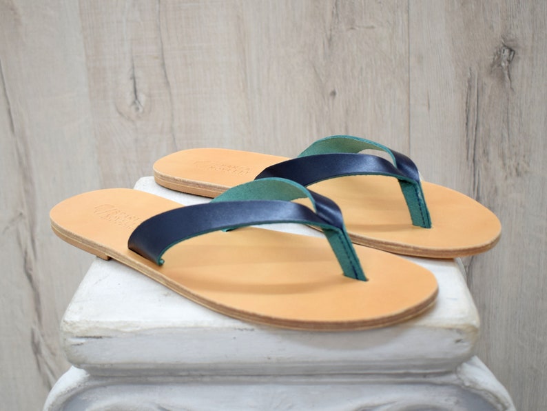 Peut inclure: Une paire de tongs en cuir avec des lani&egrave;res bleu marine et des lani&egrave;res de thong vert d'eau. Les sandales ont une semelle marron clair et sont pr&eacute;sent&eacute;es sur une surface blanche sur un fond en bois clair. Chaussures d'&eacute;t&eacute;.