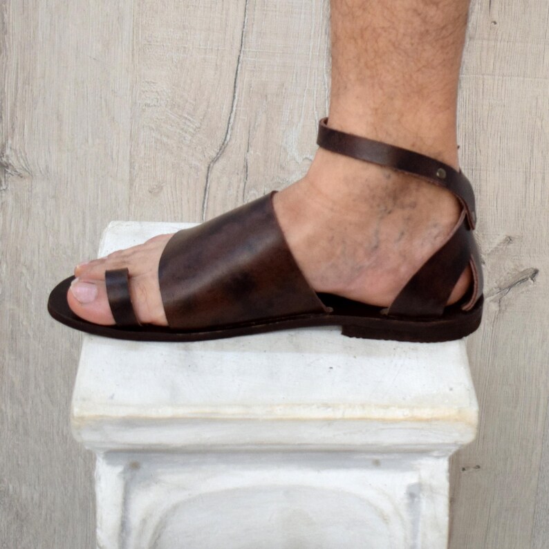 May include: Brown leather sandal with a toe loop and ankle strap. The sandal has a flat sole and a simple design. The leather appears to be smooth and the sandal is a dark brown color. The ankle strap is secured with a small metal button.