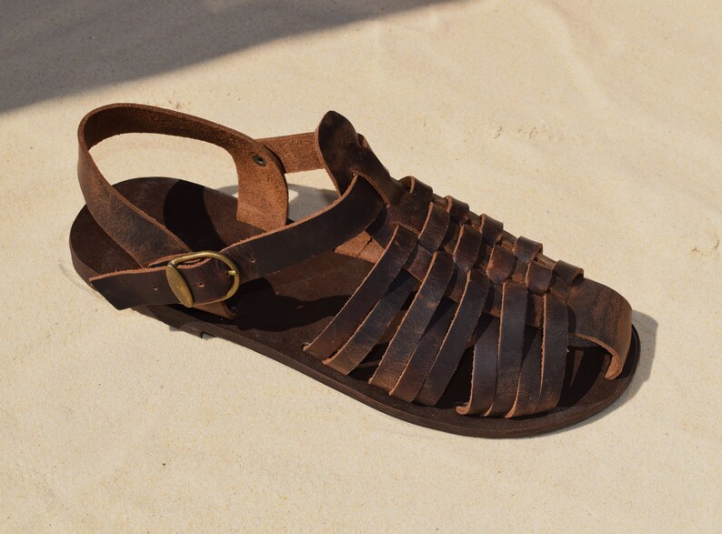 May include: A single brown leather sandal with a woven strap design. The sandal features a buckle closure and a strap around the ankle. The sandal is displayed on a sandy surface.