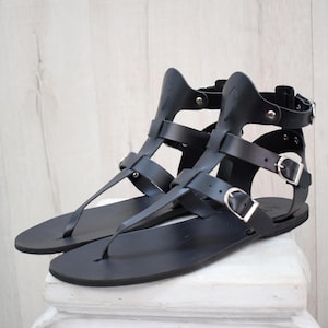 May include: A pair of black leather gladiator sandals. The sandals feature multiple straps, buckles, and a thong-style toe post. The design includes a high ankle cuff and a flat sole. The sandals are displayed on a white surface against a light wood background.