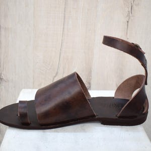 May include: Brown leather sandal with a toe loop and ankle strap. The sandal features a wide strap across the top of the foot and a buckle closure on the ankle strap. The leather has a rich, dark brown color.