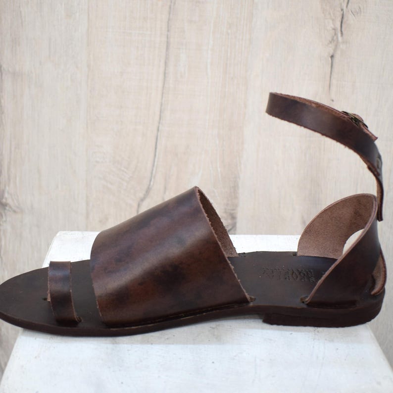 May include: Brown leather sandal with a toe loop and ankle strap. The sandal features a wide strap across the top of the foot and a buckle closure on the ankle strap. The leather has a rich, dark brown color.