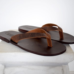Peut inclure: Une paire de tongs en cuir marron. Les sandales ont des semelles marron fonc&eacute; et des lani&egrave;res marron clair. Les tongs sont pr&eacute;sent&eacute;es sur une surface blanche sur fond blanc. Les sandales sont de style classique.