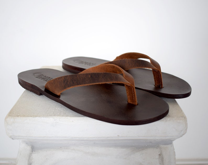 Peut inclure: Une paire de tongs en cuir marron. Les sandales ont des semelles marron fonc&eacute; et des lani&egrave;res marron clair. Les tongs sont pr&eacute;sent&eacute;es sur une surface blanche sur fond blanc. Les sandales sont de style classique.