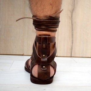 Puede incluir: Una sandalia de gladiador de cuero marr&oacute;n oscuro con suela plana, una tira vertical en el tal&oacute;n asegurada con remaches met&aacute;licos y largos cordones de cuero que se envuelven alrededor del tobillo, hecha de tiras de cuero grueso y resistente.