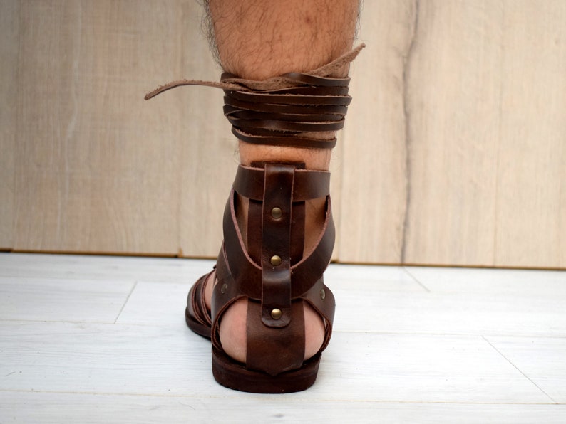 Puede incluir: Una sandalia de gladiador de cuero marr&oacute;n oscuro con suela plana, una tira vertical en el tal&oacute;n asegurada con remaches met&aacute;licos y largos cordones de cuero que se envuelven alrededor del tobillo, hecha de tiras de cuero grueso y resistente.