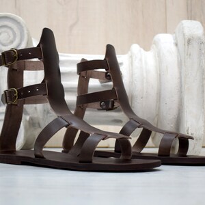 May include: A pair of brown leather gladiator sandals with multiple straps and buckle closures. The sandals have a flat sole and a high back, and are displayed on a white surface. The sandals are a classic style, suitable for warm weather.
