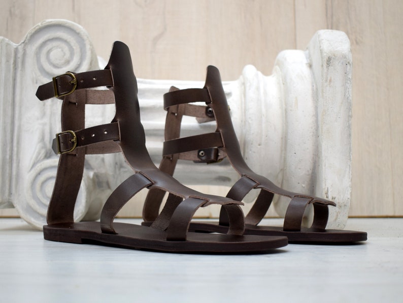 May include: A pair of brown leather gladiator sandals with multiple straps and buckle closures. The sandals have a flat sole and a high back, and are displayed on a white surface. The sandals are a classic style, suitable for warm weather.