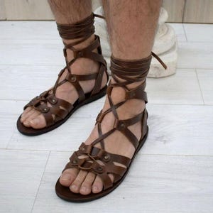 Puede incluir: Sandalias de gladiador en cuero marr&oacute;n oscuro con m&uacute;ltiples tiras que envuelven el pie y suben hasta el tobillo. El dise&ntilde;o incluye remaches met&aacute;licos que aseguran las bandas de cuero entrecruzadas, una suela plana y cordones largos para atar.