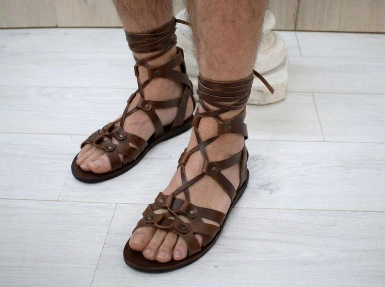 Puede incluir: Sandalias de gladiador en cuero marr&oacute;n oscuro con m&uacute;ltiples tiras que envuelven el pie y suben hasta el tobillo. El dise&ntilde;o incluye remaches met&aacute;licos que aseguran las bandas de cuero entrecruzadas, una suela plana y cordones largos para atar.