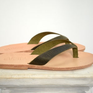 Peut inclure: Paire de tongs en cuir avec des lani&egrave;res vert olive. Les sandales ont une semelle marron clair. Les lani&egrave;res sont textur&eacute;es et les sandales semblent faites &agrave; la main. Elles sont pr&eacute;sent&eacute;es sur une surface blanche.