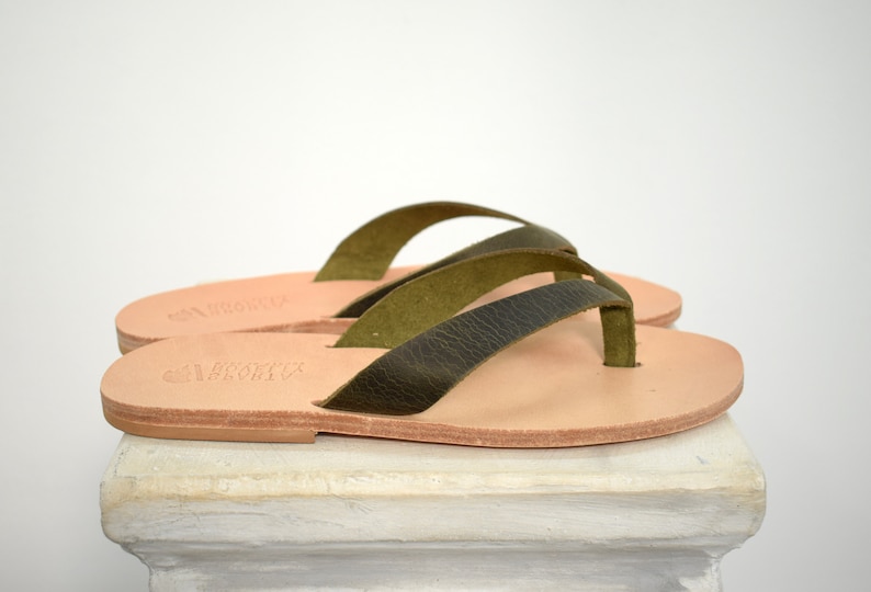 Peut inclure: Paire de tongs en cuir avec des lani&egrave;res vert olive. Les sandales ont une semelle marron clair. Les lani&egrave;res sont textur&eacute;es et les sandales semblent faites &agrave; la main. Elles sont pr&eacute;sent&eacute;es sur une surface blanche.