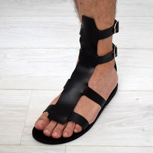 May include: Black leather gladiator sandals with multiple straps and buckle closures. The sandals feature a flat sole and a design that extends up the ankle. The footwear is designed to be worn on the foot.