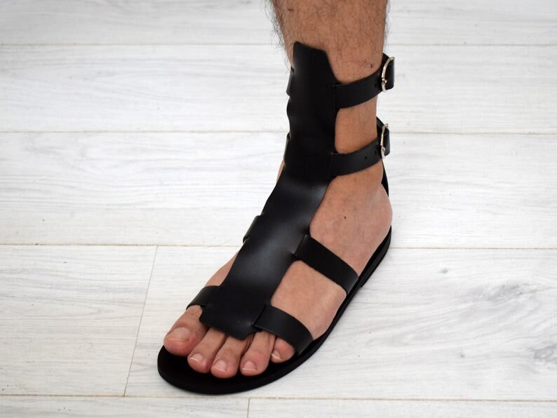 May include: Black leather gladiator sandals with multiple straps and buckle closures. The sandals feature a flat sole and a design that extends up the ankle. The footwear is designed to be worn on the foot.