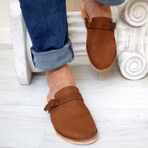 May include: Brown leather slip-on sandals with a buckle strap. The sandals have a textured appearance and a light-colored sole. The image shows the sandals being worn with blue jeans.