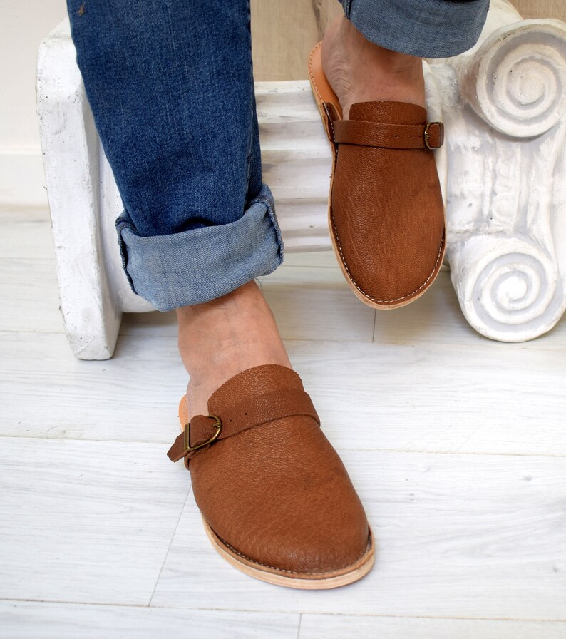 May include: Brown leather slip-on sandals with a buckle strap. The sandals have a textured appearance and a light-colored sole. The image shows the sandals being worn with blue jeans.
