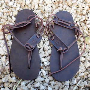 May include: A pair of brown leather sandals with laces that wrap around the ankles. The sandals have a simple design and are made of high-quality leather.
