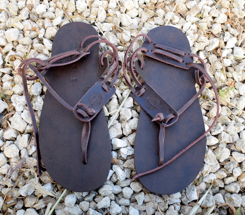May include: A pair of brown leather sandals with laces that wrap around the ankles. The sandals have a simple design and are made of high-quality leather.
