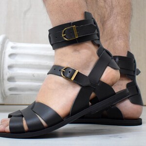May include: Black leather gladiator sandals with gold-colored buckles. The sandals feature multiple straps and a closed heel design. The ankle straps are adjustable. The sandals are designed for comfort and style.