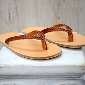 Op de afbeelding: Een paar lichtbruine leren teenslippers. De sandalen hebben een gevlochten band en een lichtbruine zool. De teenslippers worden gepresenteerd op een wit oppervlak tegen een lichte houten achtergrond. De sandalen zijn neutraal van kleur.
