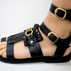 May include: Black leather gladiator sandals with gold-colored buckles and decorative accents. The sandals feature a fringed toe strap and a lion's head detail. The ankle straps have adjustable buckles for a secure fit. The sandals are designed for a stylish, open-toe look.