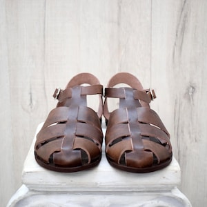 May include: A pair of brown leather sandals with a t-strap design. The sandals have a buckle closure and a rounded toe. The shoes are displayed on a white surface against a light wood background.