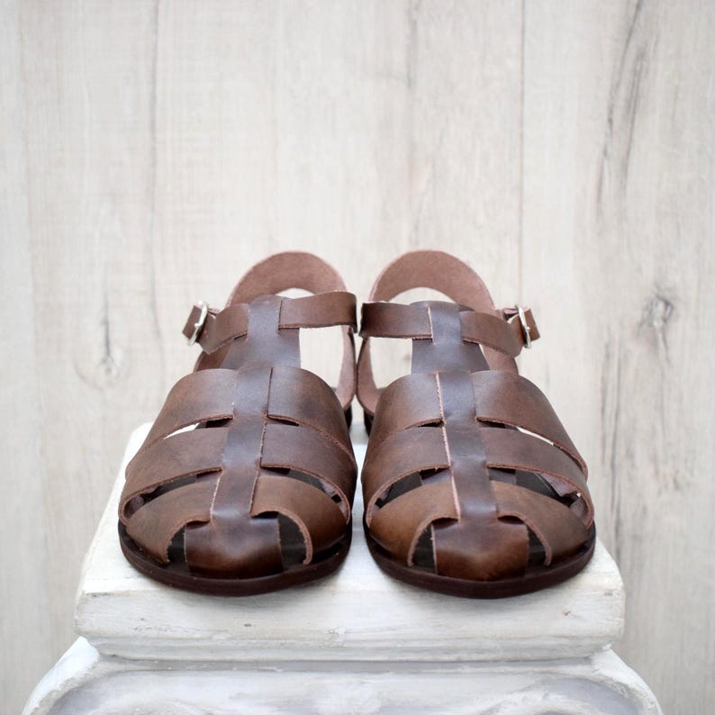 May include: A pair of brown leather sandals with a t-strap design. The sandals have a buckle closure and a rounded toe. The shoes are displayed on a white surface against a light wood background.