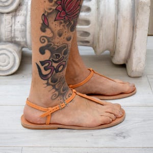 May include: A pair of orange leather sandals with a T-strap design. The sandals have a buckle closure and are worn on a light wooden floor. The image also shows a leg with a tattoo.