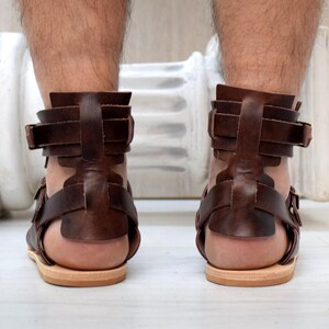 May include: Brown leather sandals with multiple straps and buckles. The sandals have a flat sole and are designed to cover the foot and ankle. The image shows the back of the sandals, highlighting the intricate strap design.