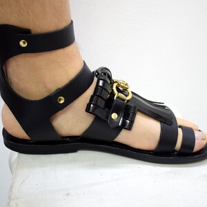 May include: Black leather gladiator sandal with gold-colored hardware. The sandal features an ankle strap, multiple straps across the foot, and decorative elements. The design includes a fringe detail and a decorative buckle.