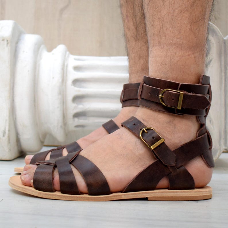 May include: Brown leather sandals with multiple straps and buckle closures. The sandals have a flat sole and are designed with an open toe. The ankle straps feature a brass buckle. The sandals are a classic style.