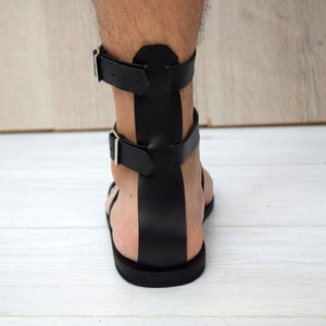 May include: Black leather gladiator sandals with two adjustable buckle straps. The sandals have a flat sole and a design that wraps around the ankle. The sandals are shown from the back view.