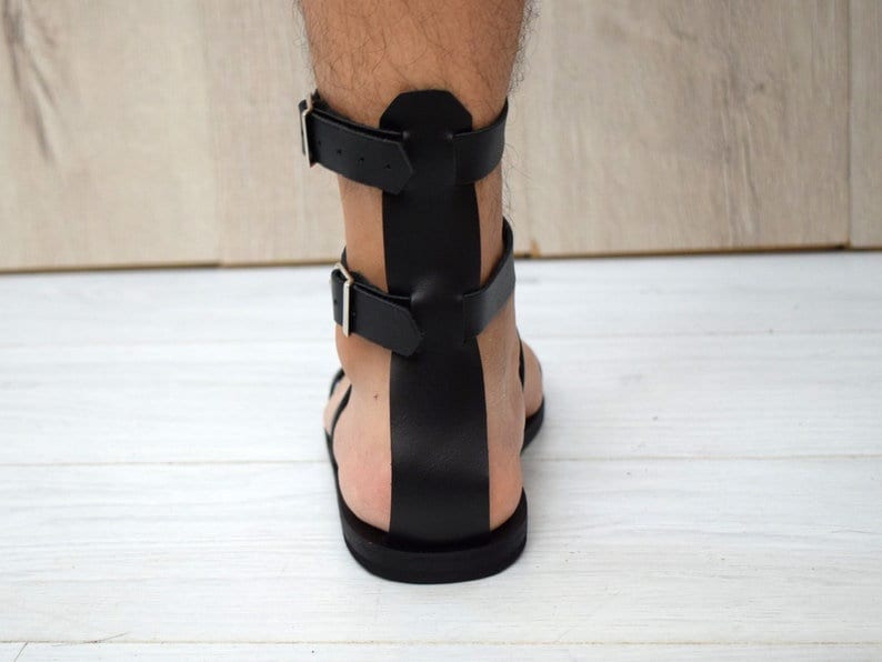 May include: Black leather gladiator sandals with two adjustable buckle straps. The sandals have a flat sole and a design that wraps around the ankle. The sandals are shown from the back view.