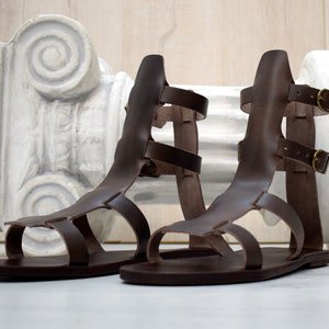 May include: A pair of brown leather gladiator sandals with multiple straps and gold-colored buckles. The sandals have a flat sole and are displayed in front of a white, classical-style column.