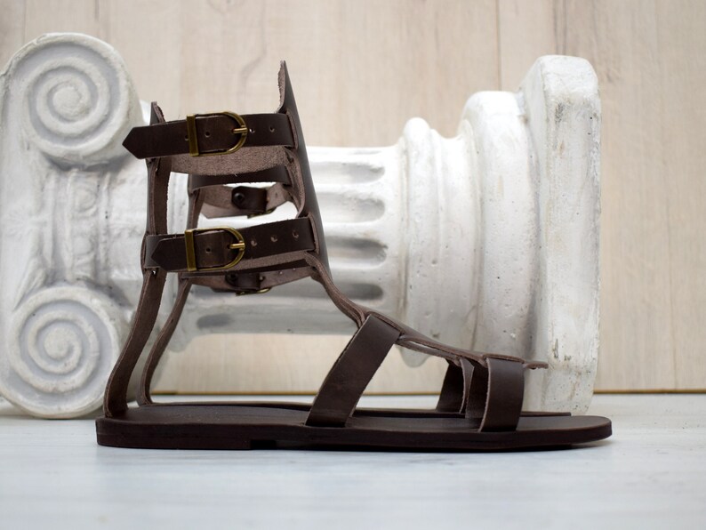 May include: A brown leather gladiator sandal with multiple straps and brass buckles. The sandal has a flat sole and is designed with an open toe. The background features a white architectural column.