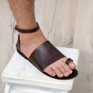 May include: Brown leather sandal with a toe loop and ankle strap. The sandal features a wide strap across the top of the foot and a buckle closure on the ankle strap. The sole is dark brown.