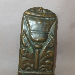 May include: A bronze decorative plaque with a raised design of a stylized flower with leaves. The plaque has a rectangular shape and a smooth surface.