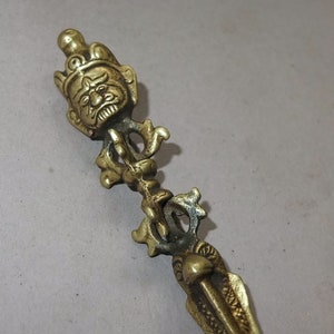 Könnte beinhalten: Ein ritueller Phurba-Dolch aus Messing mit einem detaillierten Gottheitskopf an der Spitze, einem zentralen Griff mit kunstvollen, geschwungenen Metallarbeiten und einer scharfen, spitzen Klinge am unteren Ende. Das Metall hat ein gealtertes Finish.