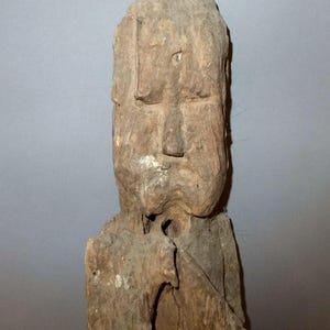 May include: A weathered, carved wooden sculpture of a human figure. The sculpture has a rectangular head with a prominent nose and a simple, blocky form. The wood is a light brown color with visible grain and some areas of discoloration.