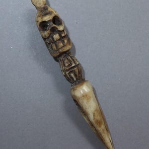Peut inclure: Un poignard en os sculpté avec une poignée en forme de crâne. Le poignard a une lame pointue et un design détaillé et texturé. La couleur est beige clair avec des accents bruns plus foncés.