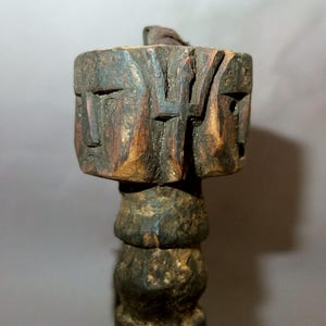 Könnte beinhalten: Eine geschnitzte Holzskulptur mit zwei Gesichtern auf einem quadratischen Block, mit einem zylindrischen Sockel. Die Skulptur besteht aus dunkelbraunem Holz mit sichtbarer Maserung und geschnitzten Details. Die Gesichter haben einfache, stilisierte Merkmale.