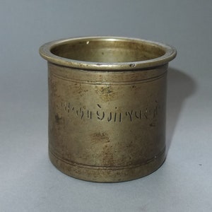 Könnte beinhalten: Ein zylindrisches Gefäß aus Messing mit einer verwitterten, metallischen Oberfläche und einem breiten, flachen Rand. Die Außenseite weist eine Reihe horizontaler Gravurlinien und ein zentrales Band mit geätzten, nicht-lateinischen Schriftzeichen auf.