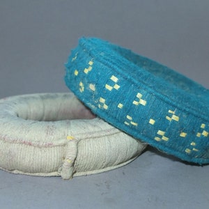 以下が含まれることがあります： 2つの円形のパッド入りオブジェクト。1つは薄いクリーム色で、もう1つは黄色の正方形のアクセントが付いたターコイズ色です。オブジェクトは布でできており、おそらくサポートまたはクッションに使用されます。ターコイズ色のオブジェクトはクリーム色のオブジェクトの上にあります。