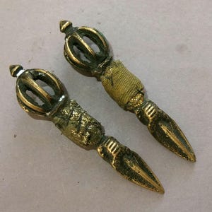 Könnte beinhalten: Zwei antike Vajra-Zepter aus Messing. Jedes Zepter hat eine detaillierte kronenartige Spitze, einen strukturierten Griff und ein spitzes Ende. Das Messing hat eine warme, gealterte Patina, die auf eine historische Verwendung hindeutet. Diese zeremoniellen Objekte sind wahrscheinlich östlichen Ursprungs.