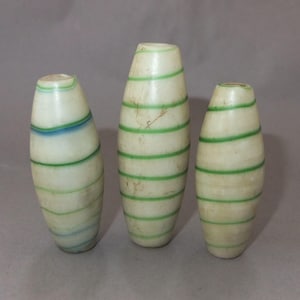 Pode incluir: Três objetos decorativos ovais brancos com listras verdes horizontais. Um objeto tem listras azuis. Os objetos variam em tamanho e são exibidos em um fundo neutro. Os objetos parecem ser feitos de um material liso.