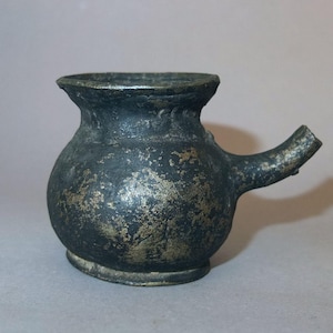 Pode incluir: Jarro de metal antigo e pequeno com corpo arredondado, bico e alça. O jarro tem uma superfície escura e texturizada com detalhes dourados. A borda do jarro é ligeiramente alargada.