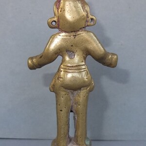 May include: A small, gold-toned brass figurine of a standing figure with arms extended outward. The figure features a pointed headdress, circular ear ornaments, and a simple garment around the waist, all mounted on a square, tiered base.