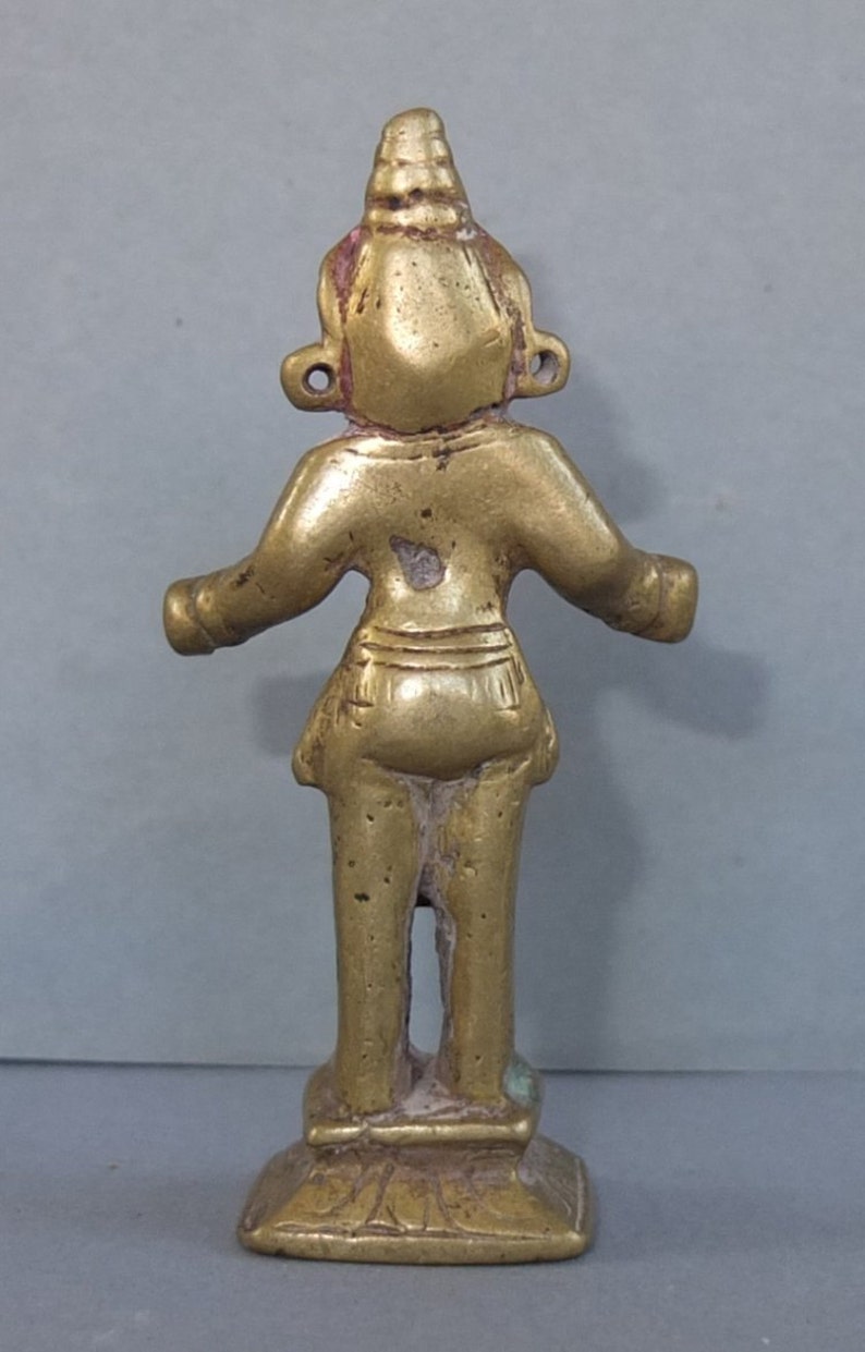 May include: A small, gold-toned brass figurine of a standing figure with arms extended outward. The figure features a pointed headdress, circular ear ornaments, and a simple garment around the waist, all mounted on a square, tiered base.