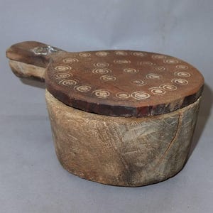 Könnte beinhalten: Ein Holzbehälter mit Deckel. Der Deckel ist mit einem Kreis-Muster verziert. Der Behälter ist aus rauhm Holz gefertigt und hat eine natürliche braune Farbe.